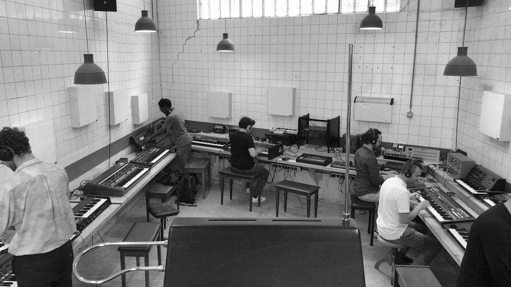 Photo en noir et blanc d'une pièce carrelée. Sept personnes se penchent sur différents instruments de musique électroniques.