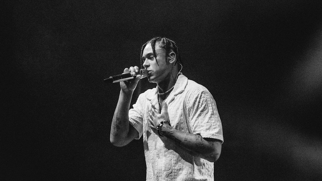 A live shot of Pronto performing. He holds a microphone with his right hand, the left hand resting on his chest.