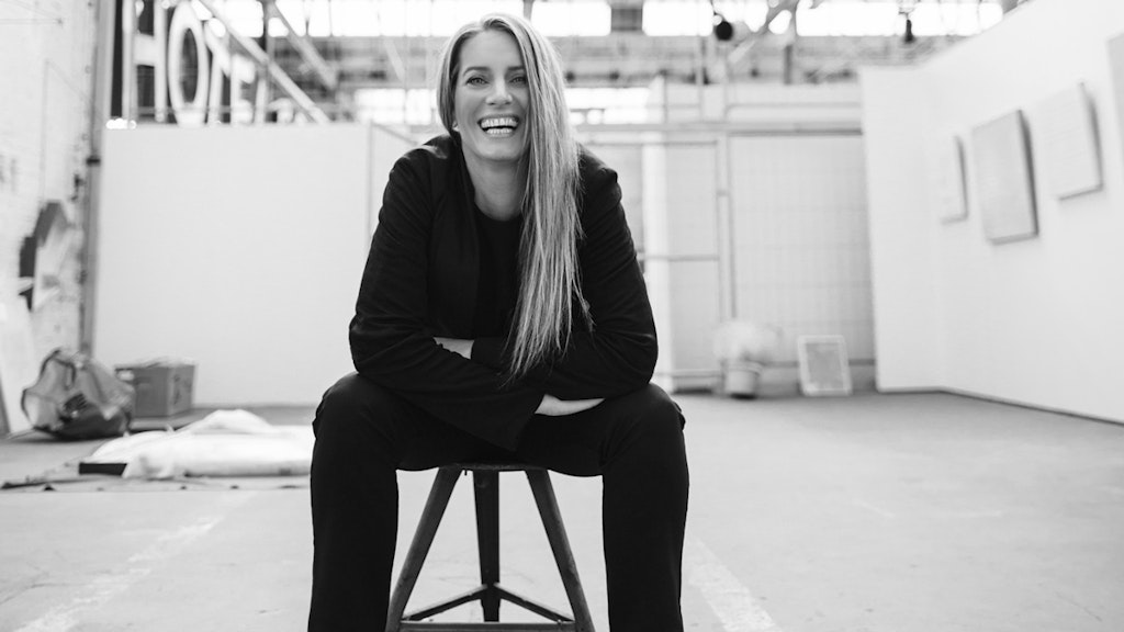 Portrait de Sonja Moonear. Elle est assise sur un tabouret, détendue, penchée en avant et rit un grand rire à la caméra.