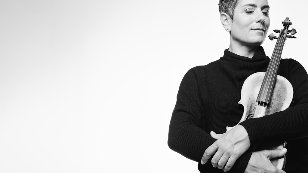 Black and white photo of Leila Schayegh. She holds her violin in front of her chest with her eyes closed.