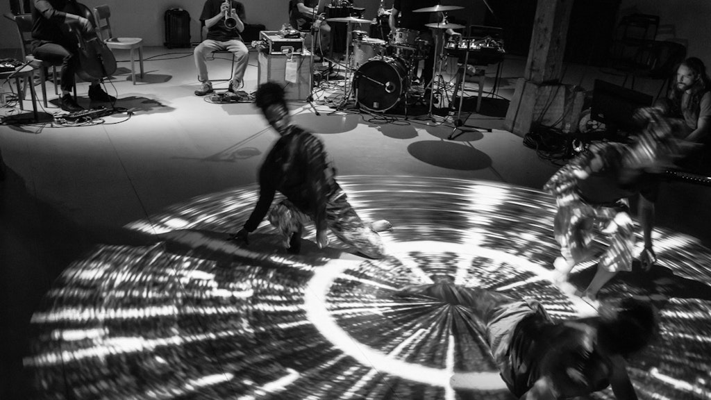 In the background, four musicians sit on chairs, playing their instruments. In the foreground, three female performers dance in a ray of light.