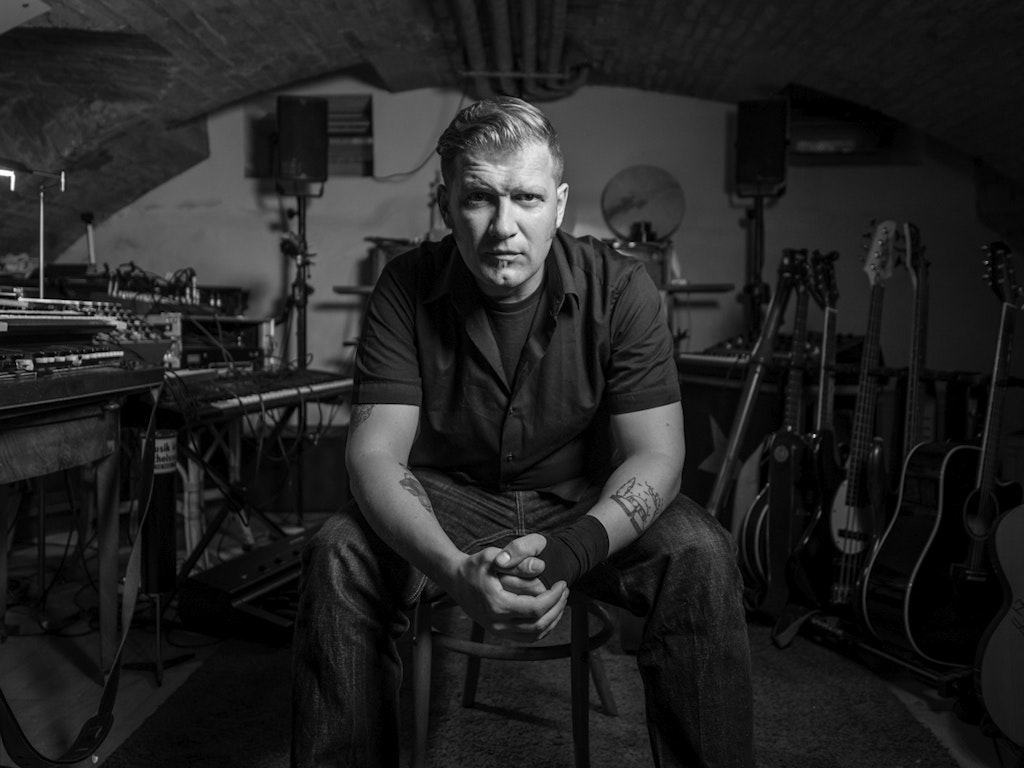Portrait of Mario Batkociv sitting on a chair, looking directly into the camera. In the background a multitude of different instruments.