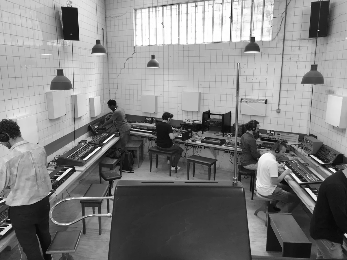 Black and white photograph of a tiled room. Seven people hunched over various electronic musical instruments.