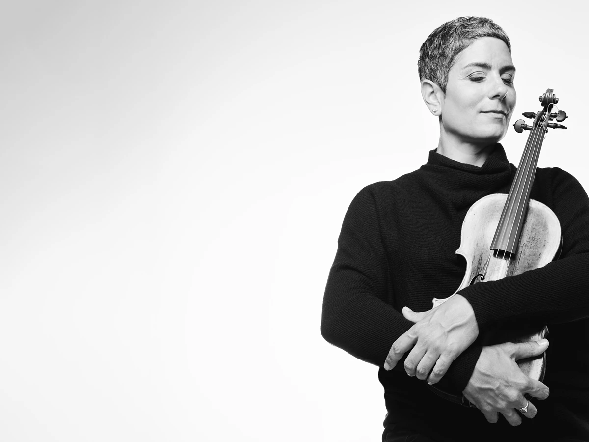 Black and white photo of Leila Schayegh. She holds her violin in front of her chest with her eyes closed.