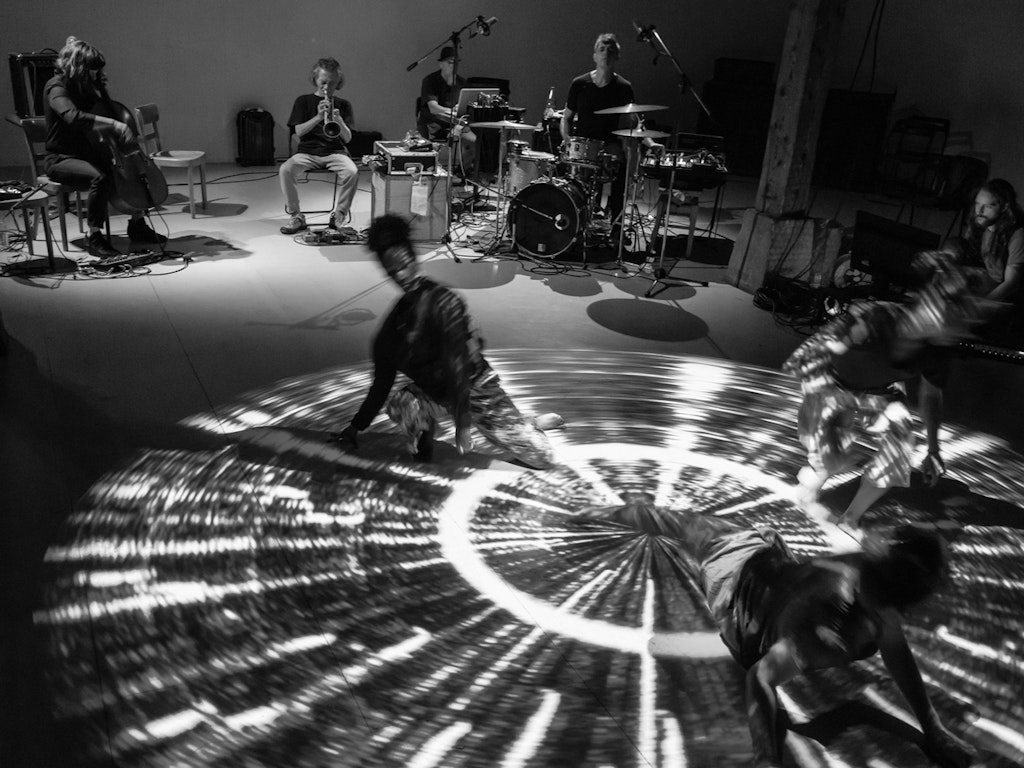 In the background, four musicians sit on chairs, playing their instruments. In the foreground, three female performers dance in a ray of light.