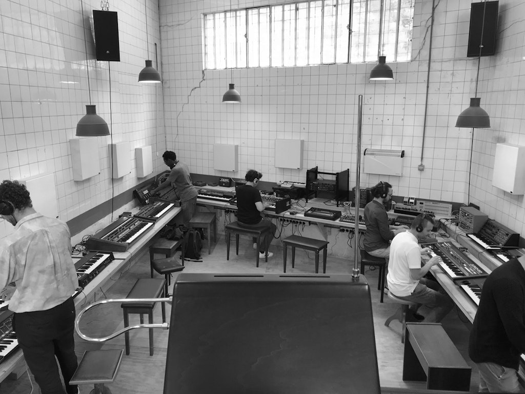 Photo en noir et blanc d'une pièce carrelée. Sept personnes se penchent sur différents instruments de musique électroniques.