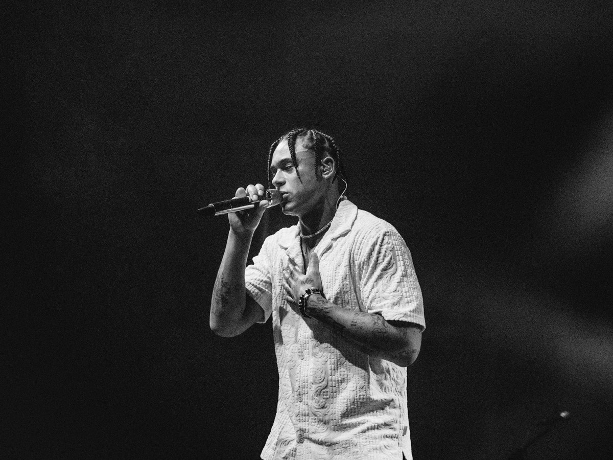 Une photo de Pronto en train de se produire en live. Il tient un microphone de la main droite, la main gauche repose sur sa poitrine.